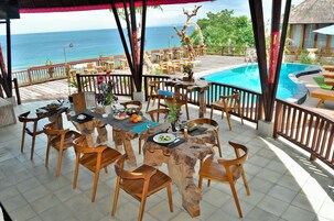 A restaurant with wooden tables and chairs, a pool, and a view of the ocean.
