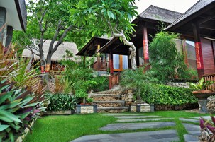 Una casa de madera tradicional con techo de paja, rodeada de abundante vegetación y una escalera de piedra que conduce a una terraza de madera.