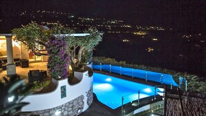 Une vue nocturne d’une piscine, avec un mur en pierre et une pergola ornée de fleurs violettes.