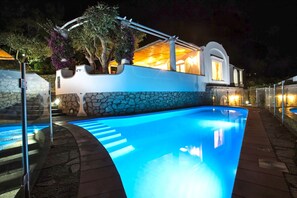 En swimmingpool med stenkant og glasafskærmning, omgivet af en stenvæg og en hvid bygning med balkon.