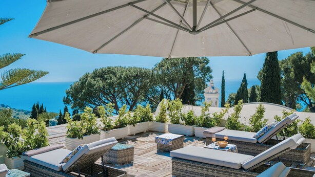 Un espace extérieur ensoleillé, aménagé avec des fauteuils confortables et un grand parasol, offrant une vue panoramique sur la mer et les arbres.