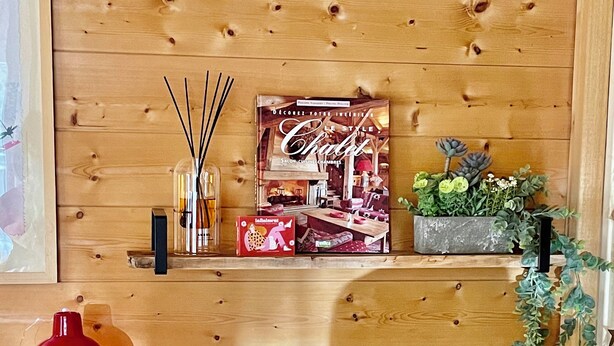 A wooden wall with shelves holding a book, a red bottle, and a potted plant. - Chalet-style apartment—3 rooms, 2 bedrooms, ground floor, small garden, parking—3 minutes from the base of the slopes, near the center (La Clusaz)
