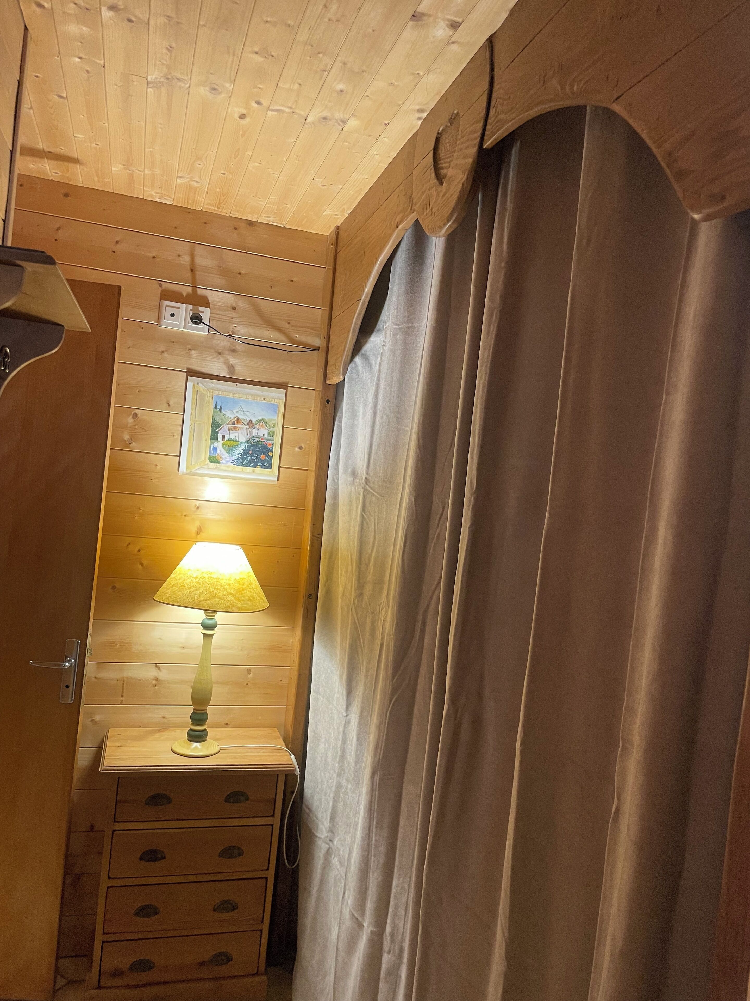 L’intérieur d’un chalet en bois, avec une commode en bois, une lampe, un tableau encadré accroché au mur et un rideau.