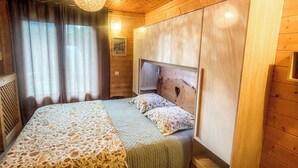 A wooden-paneled bedroom with a bed, curtains, a hanging light fixture, and a wooden wardrobe. - Chalet-style apartment—3 rooms, 2 bedrooms, ground floor, small garden, parking—3 minutes from the base of the slopes, near the center (La Clusaz)