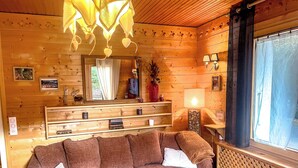 A cozy cabin interior with a wooden sofa, a mirror, a fireplace, and a window with curtains. - Chalet-style apartment—3 rooms, 2 bedrooms, ground floor, small garden, parking—3 minutes from the base of the slopes, near the center (La Clusaz)