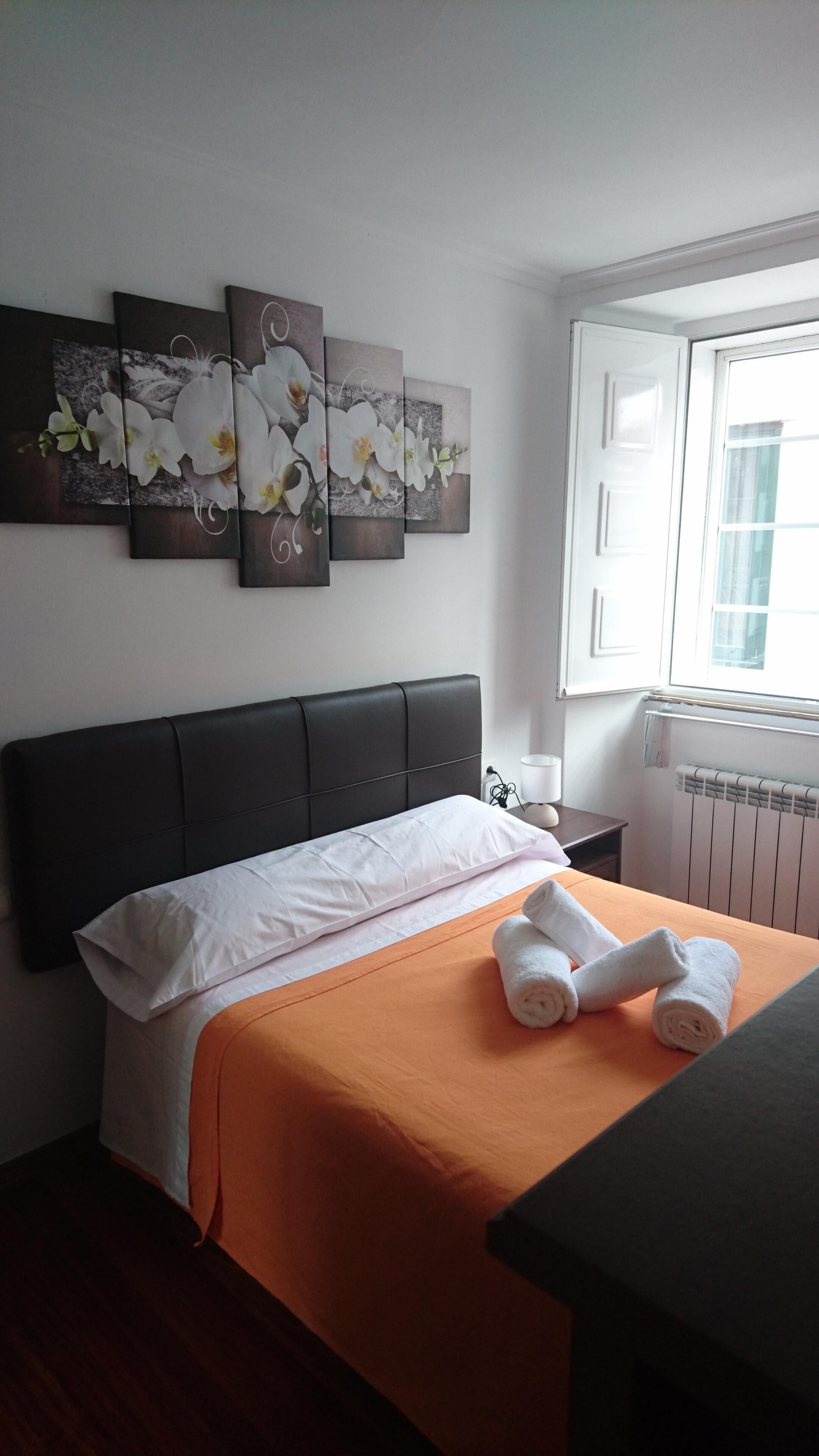 Habitación de hotel con cama, colcha naranja, almohadas blancas y cabecera. Se ven tres cuadros en la pared con flores blancas. Una ventana con persianas y un radiador en la pared.