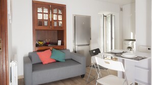 Un espace de vie compact comprenant un canapé gris, une petite table à manger, une chaise blanche, un meuble encastré en bois et un réfrigérateur moderne.