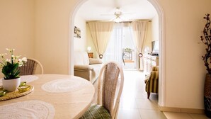 A dining area with a round table, chairs, and a potted plant. There is a doorway leading to a living area with a sofa and a television. - El Veril Duque, 2 bed apartment, sleeps 4, only 7 min walk to El Duque beach (Costa Adeje)