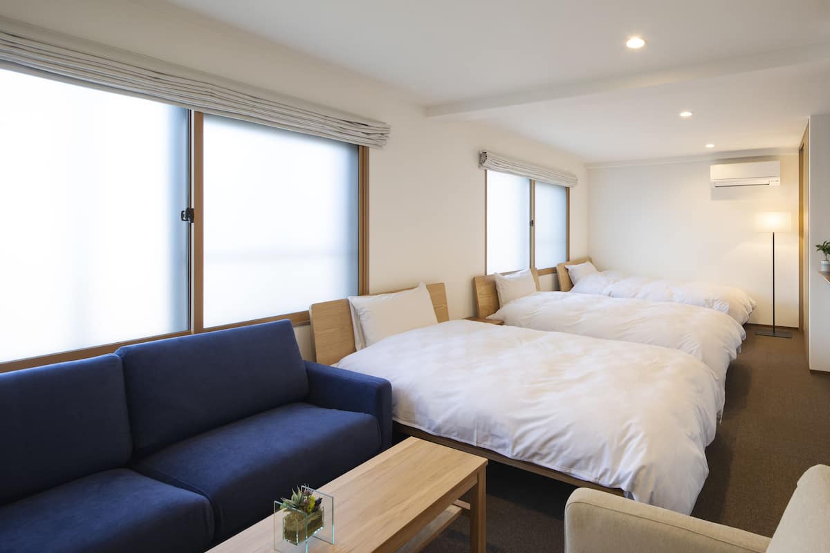 Habitación de hotel con dos camas, un sofá, una mesa pequeña y ventanas amplias.