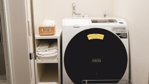A compact, white washing machine with a black drum, placed next to a shelf with folded towels and a wicker basket. - So Kyoto Fushimi Inari (Kyoto)