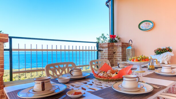 A table set for breakfast with a view of the sea, white chairs, and a brick wall.