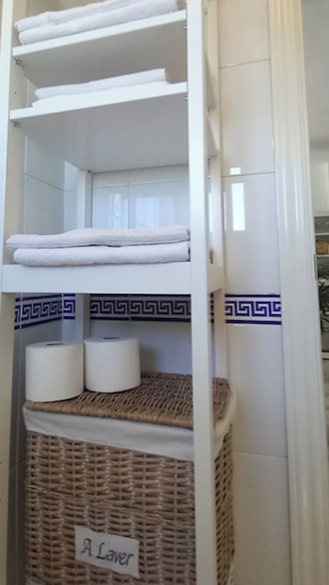 Une salle de bain avec des murs recouverts de carreaux blancs, une étagère blanche où sont posées des serviettes, un panier en osier étiqueté « À laver », et une bordure de carreaux bleus et blancs formant un motif.