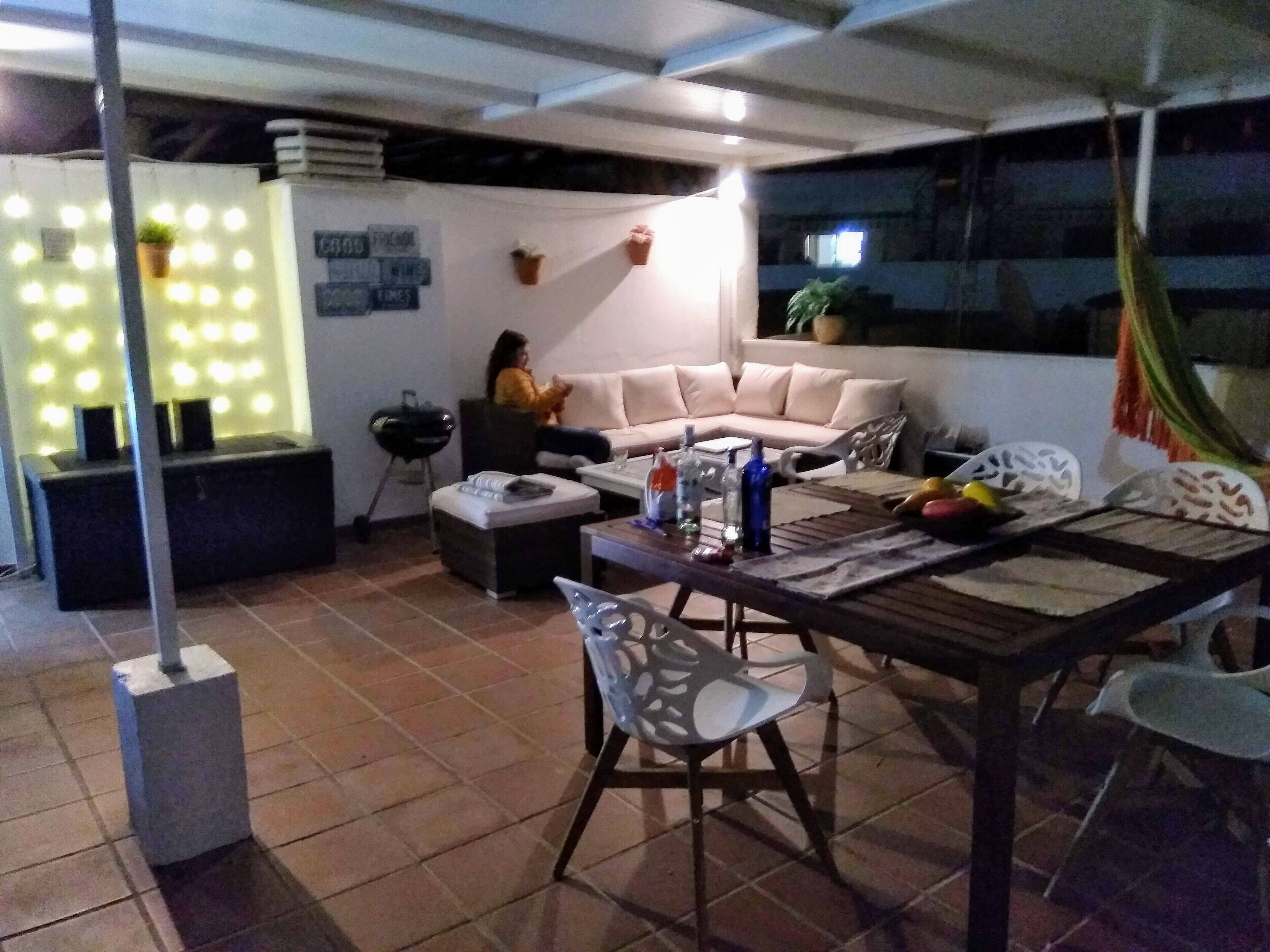 Ein Wohnbereich mit einer Couch, einem Couchtisch und einem Esstisch mit Stühlen. An der Wand stehen Topfpflanzen und in der Ecke ein Grill.