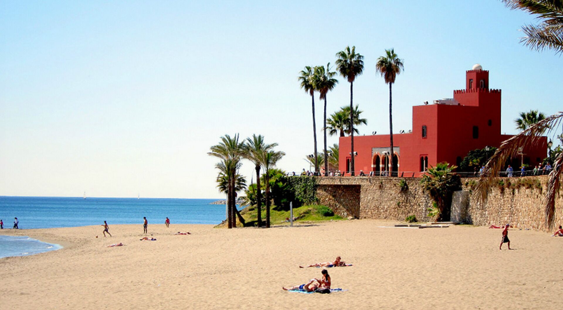 A beach with palm trees, a red building, and people sunbathing.