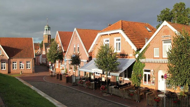 A cobblestone street lined with traditional brick buildings, some with outdoor seating and greenery. - Apartment Mühlenblick EG - Apartments Mühlenblick, 45217 (Jemgum)