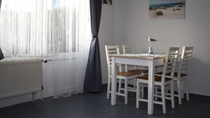 A dining area with a table and chairs, a window with curtains, a radiator, and a beach-themed picture on the wall. - Apartment Mühlenblick EG - Apartments Mühlenblick, 45217 (Jemgum)