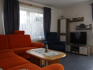 Un saló amb un sofà, una butaca i una taula petita amb un diari. Hi ha una televisió i un moble decorat amb objectes decoratius.