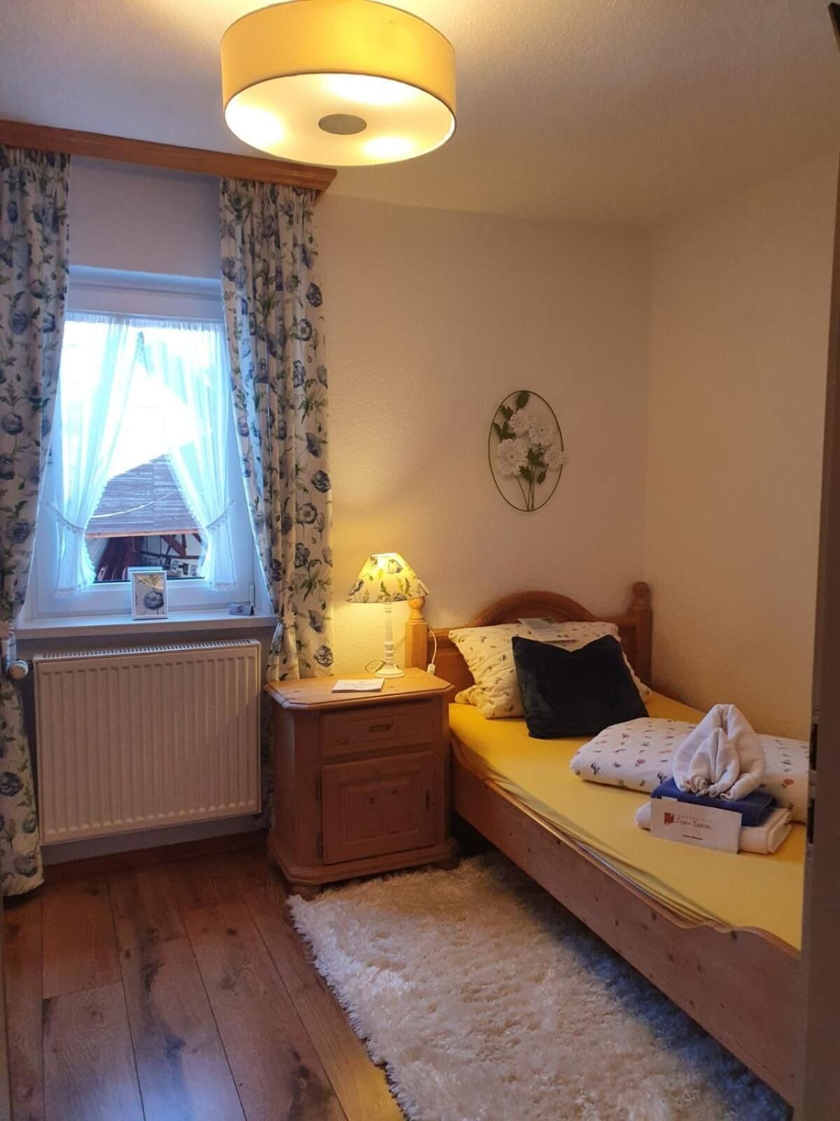 A bedroom with a bed, a nightstand, a lamp, a window with curtains, a radiator, and a wall decoration.