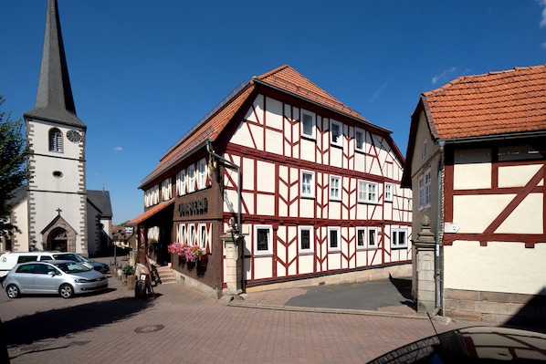 Un edificio tradicional con entramado de madera, una iglesia con un campanario alto y una calle pavimentada con coches estacionados.