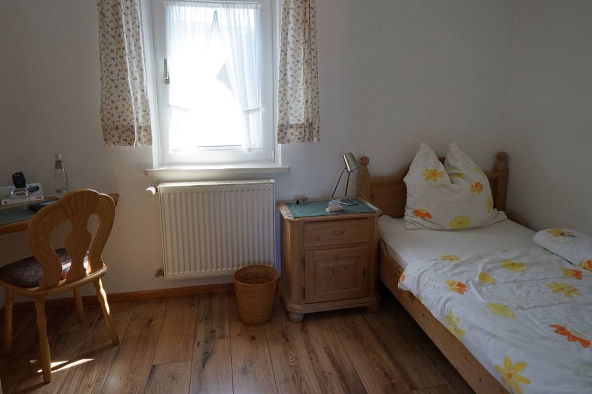 A bedroom with a wooden floor, a bed with floral bedding, a nightstand, a chair, a radiator, and a window with floral curtains.