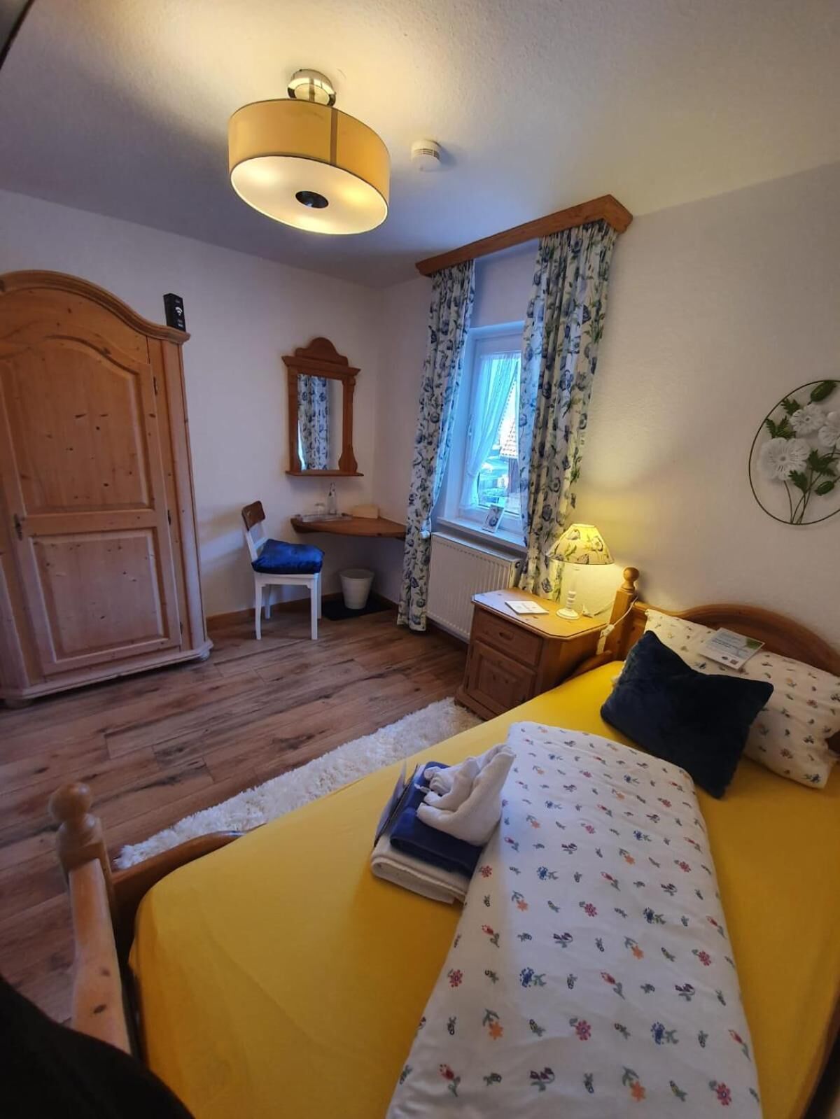 A bedroom with a bed, a desk, a wardrobe, a mirror, and a window with curtains.