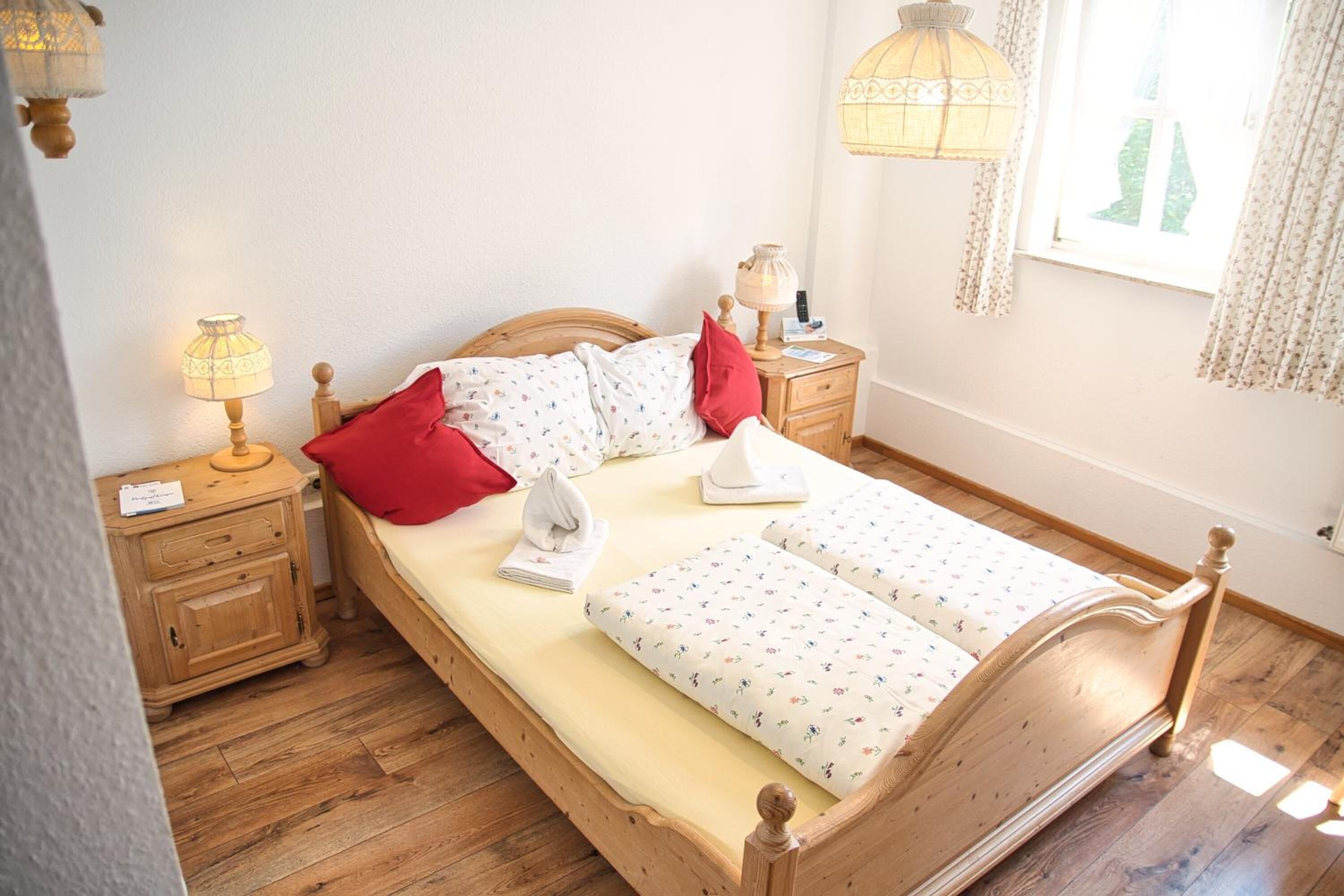 Una cama de madera con cubrecama floral, dos almohadas rojas, una toalla blanca y una mesita de noche con una lámpara.