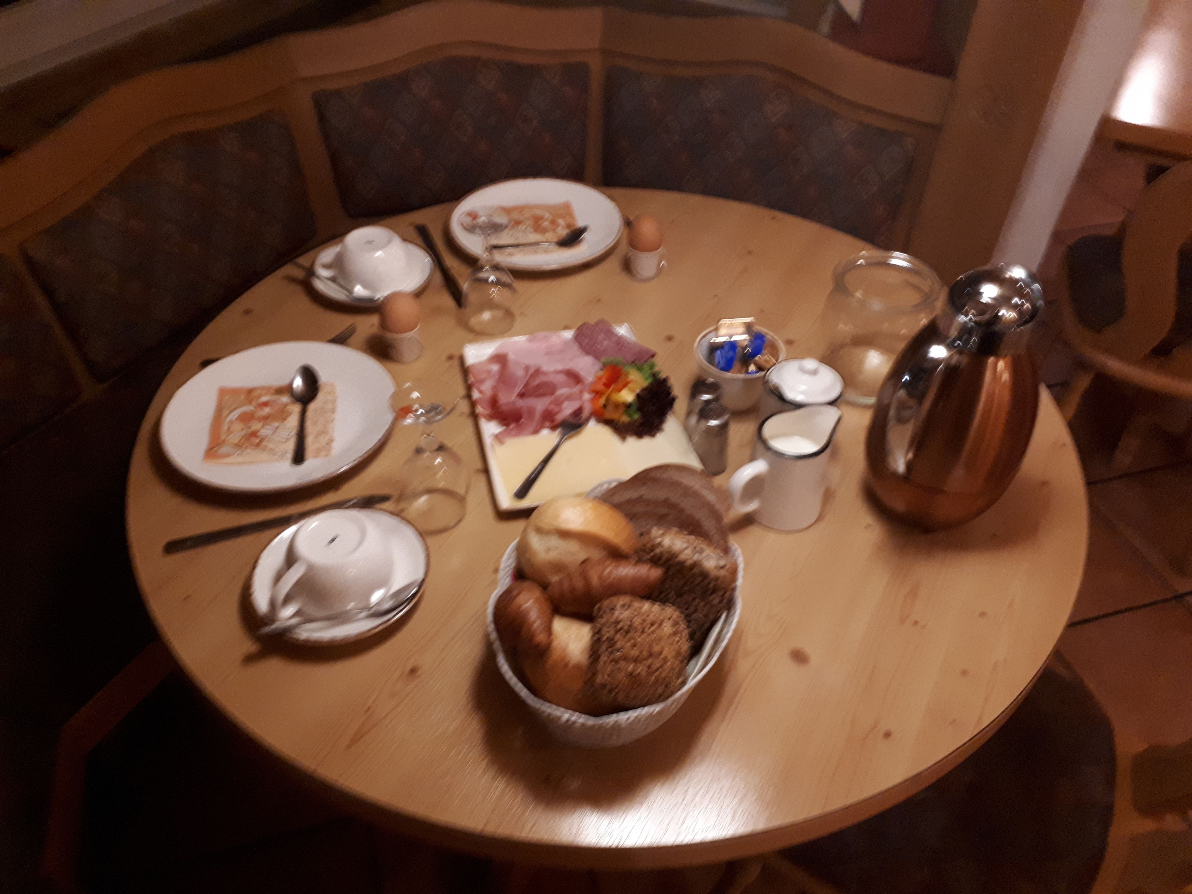 A round wooden table with plates, cutlery, a bread basket, a teapot, and a salt shaker.