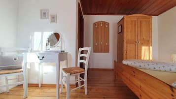Une chambre avec un plancher en bois, un bureau blanc avec un miroir, une chaise, un lit recouvert d’une couvre-lit à motifs floraux et une armoire en bois.