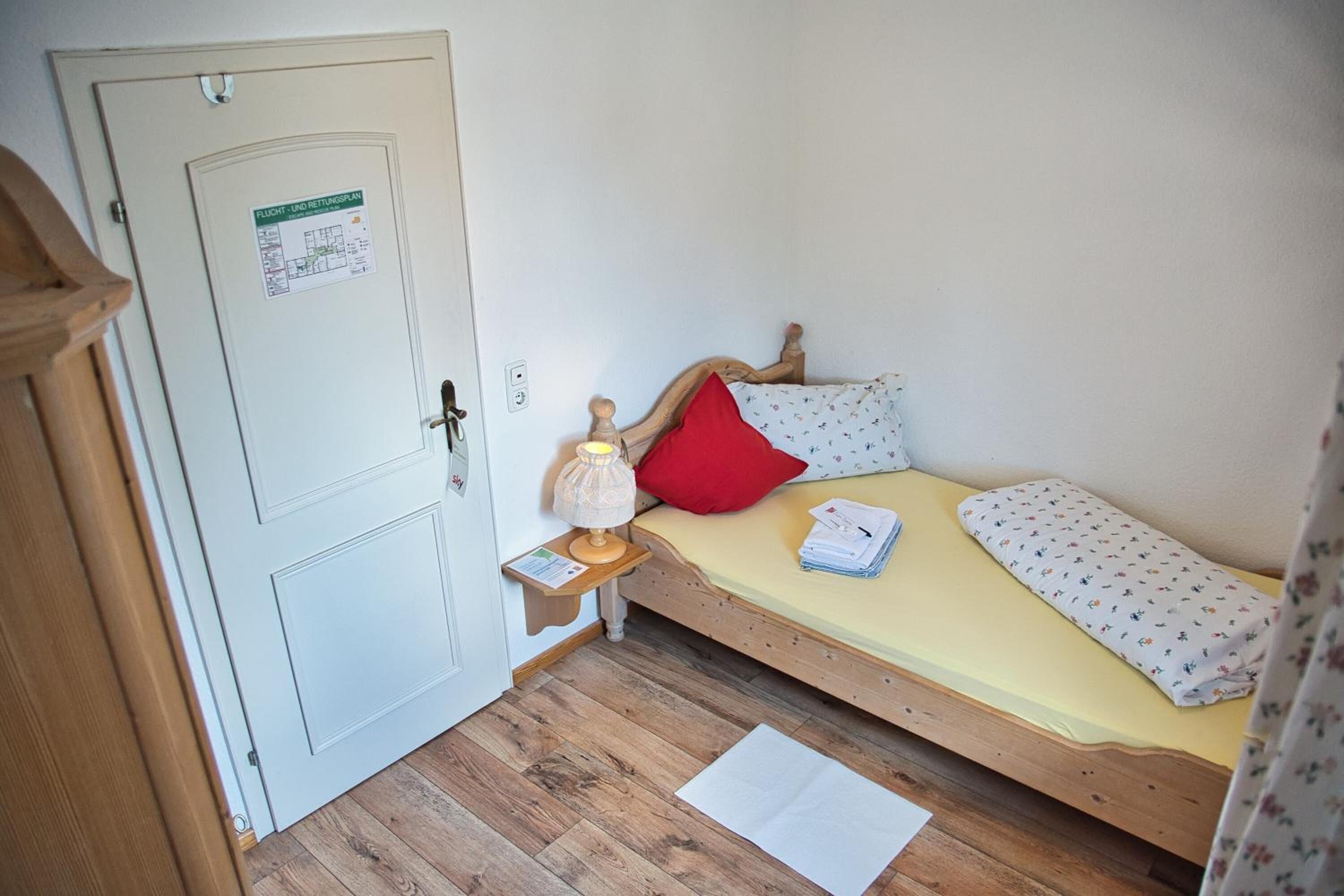 Una habitación pequeña con cama individual, suelo de madera, mesita de noche con lámpara, una puerta con un aviso y cama con almohada roja y ropa de cama estampada.