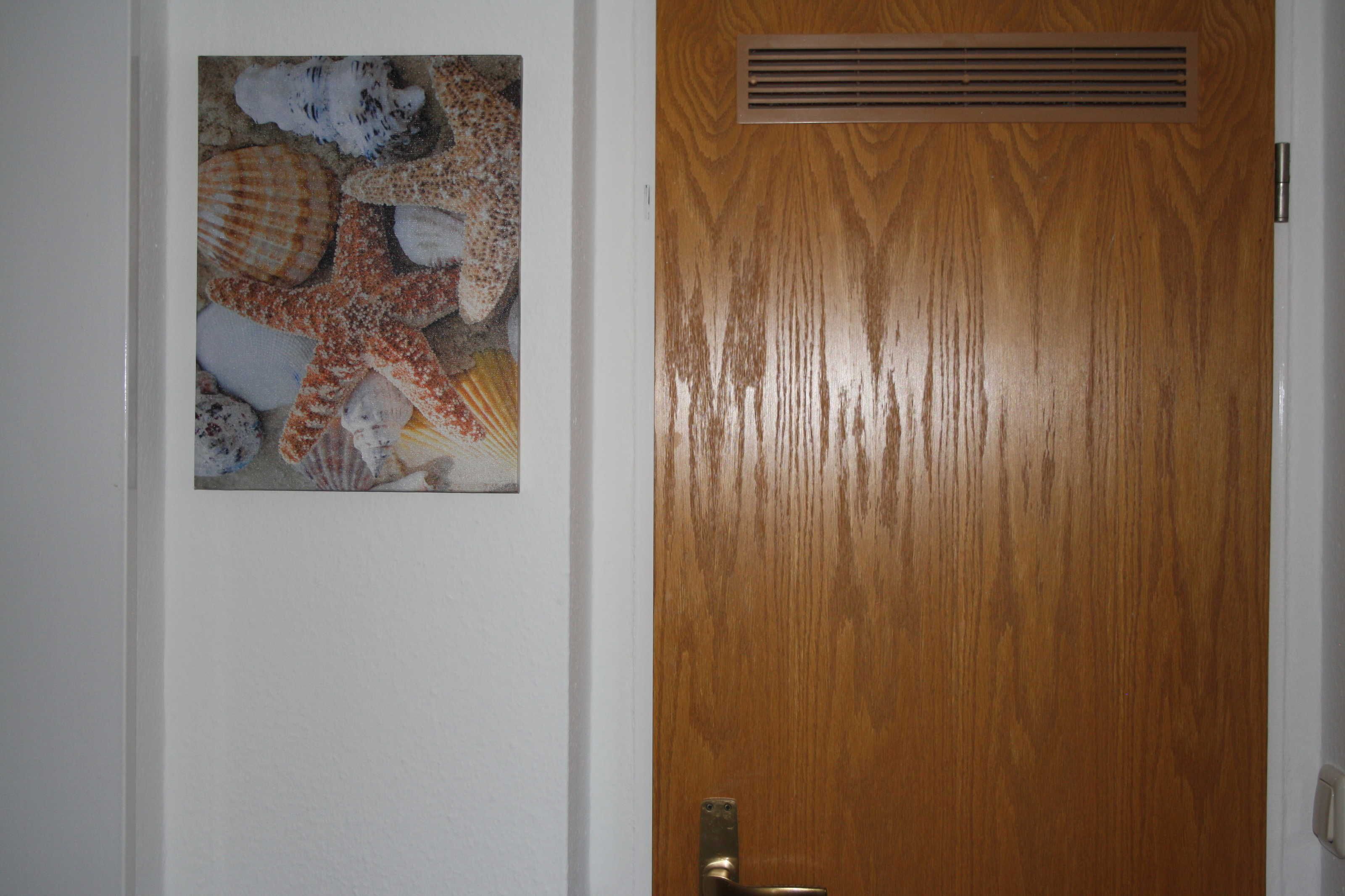 A wooden door with a vent and a framed picture of seashells on the adjacent wall.