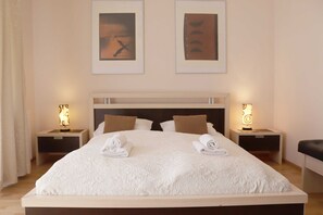 Een keurig opgemaakt bed met witte lakens en twee handdoeken, een houten hoofdbord, twee nachtkastjes met lampen, en twee ingelijste kunstwerken aan de muur.