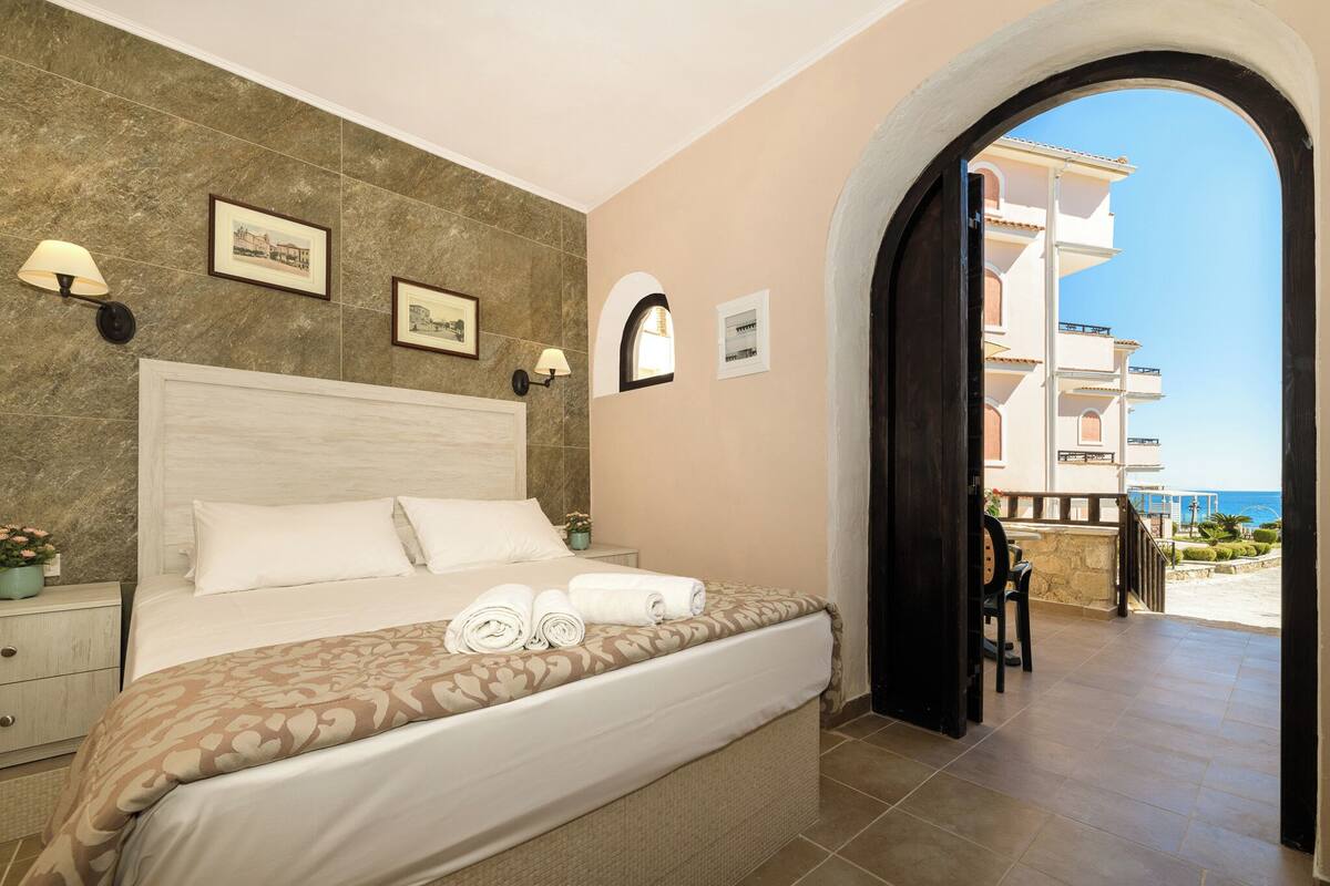 A hotel room with a large bed, a nightstand, a wall-mounted lamp, and a view of the sea through an open doorway.