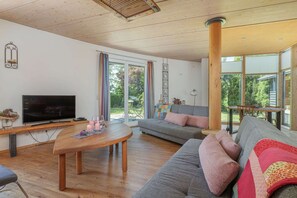 Ein modernes Wohnzimmer mit einer Couch, einem Holztisch, einem Fernseher und einem großen Fenster mit Blick auf Grünflächen.