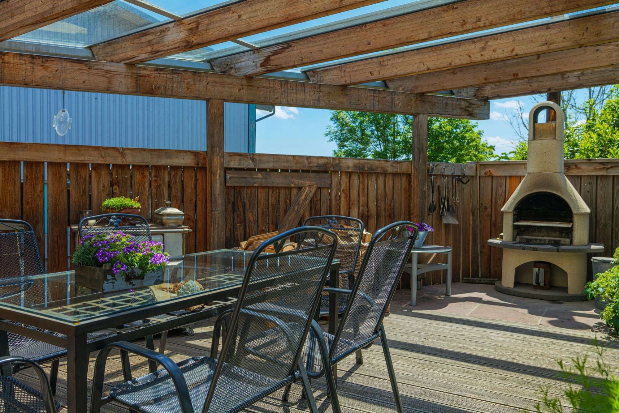 Een terras buiten met een houten pergola, een glazen tafel, metalen stoelen en een pizzaoven.