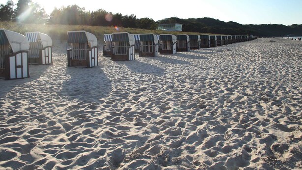 Een rij strandhutten aan het strand, met een zandstrand en een bebost heuvel in de verte.