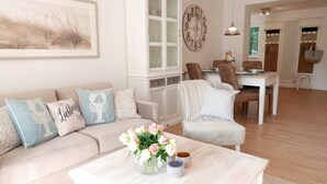 Een woonkamer met een bank, een bijzettafel met bloemen, een eethoek met stoelen en een klok aan de muur.