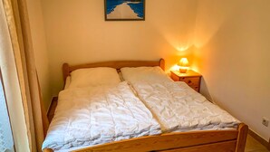 Una cama de madera individual con ropa de cama blanca, una mesita de noche con lámpara y un cuadro con una playa en la pared.
