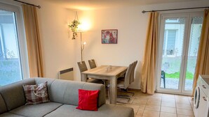 Una sala de estar con un sofá gris, una mesa de centro de madera, una mesa de comedor con sillas y una zona de cocina con lavadora.