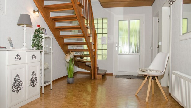 A room with a wooden staircase, a white cabinet with decorative patterns, a white chair, and a potted plant. - Vacation home Voß, 65260 - Holiday home in East Frisia, 130 sqm, 4 stars (Moormerland)