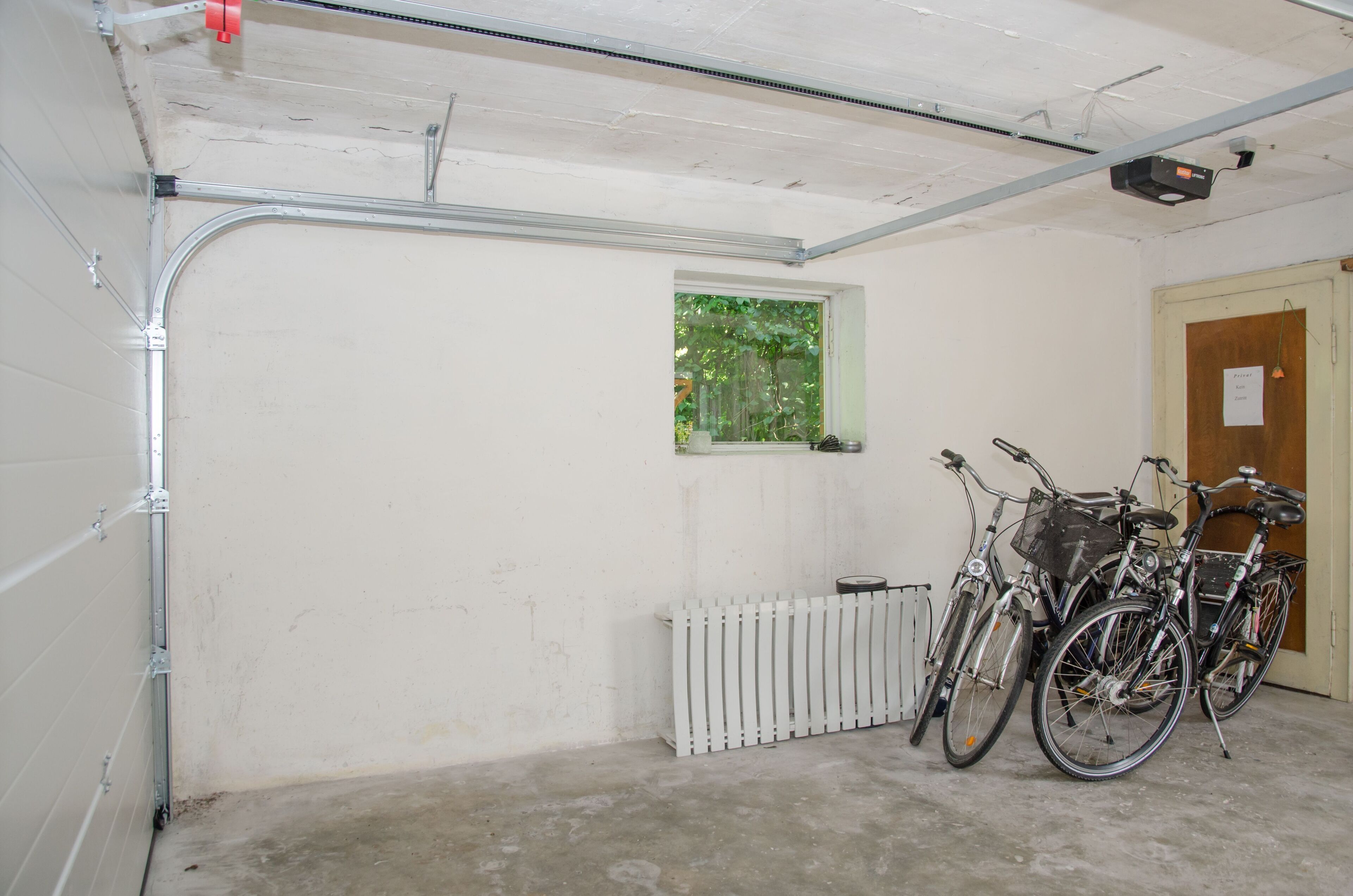 A garage with two bicycles parked, a white radiator, and a window with greenery visible outside.