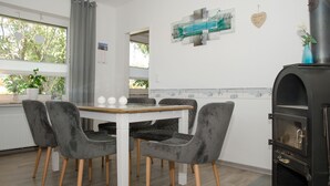 A dining area with a white table and grey upholstered chairs, a black wood-burning stove, and a window with a view of trees. - Vacation home Voß, 65260 - Holiday home in East Frisia, 130 sqm, 4 stars (Moormerland)
