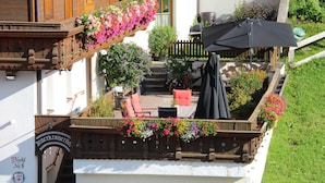 Un balcón de madera con cestas de flores colgantes, mobiliario exterior y una zona de barbacoa.