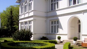 Ein weißes Gebäude mit einem repräsentativen Eingang, einem gepflasterten Weg und gepflegten Hecken.