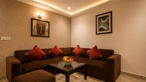 Una sala de estar con un sofá marrón, cojines rojos, una mesa de centro de vidrio con fruta y dos cuadros en la pared.