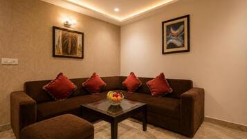 A living room with a brown sofa, red cushions, a glass coffee table with fruit, and two framed pictures on the wall.