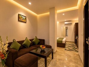 A modern hotel room with a sofa, a glass coffee table, a fruit bowl, a bed with a green bedspread, and a bathroom with a mirror and air conditioning unit. - La-Sapphire Hotel (New Delhi)