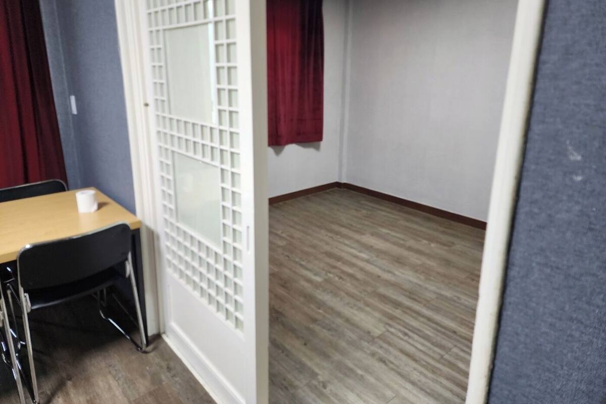 A room with a wooden floor, a white partition with a grid design, a red curtain, and a table with a chair.