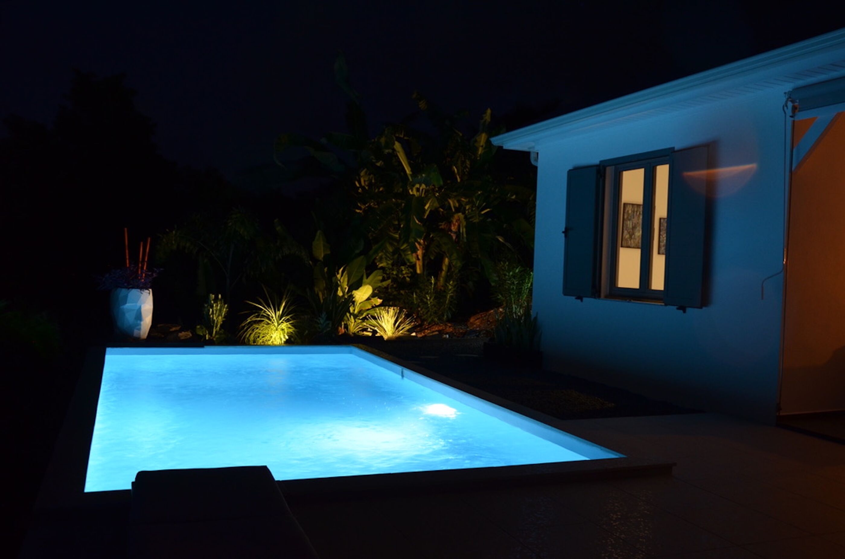 Piscina com borda iluminada, cercada por plantas e um edifício com janela.