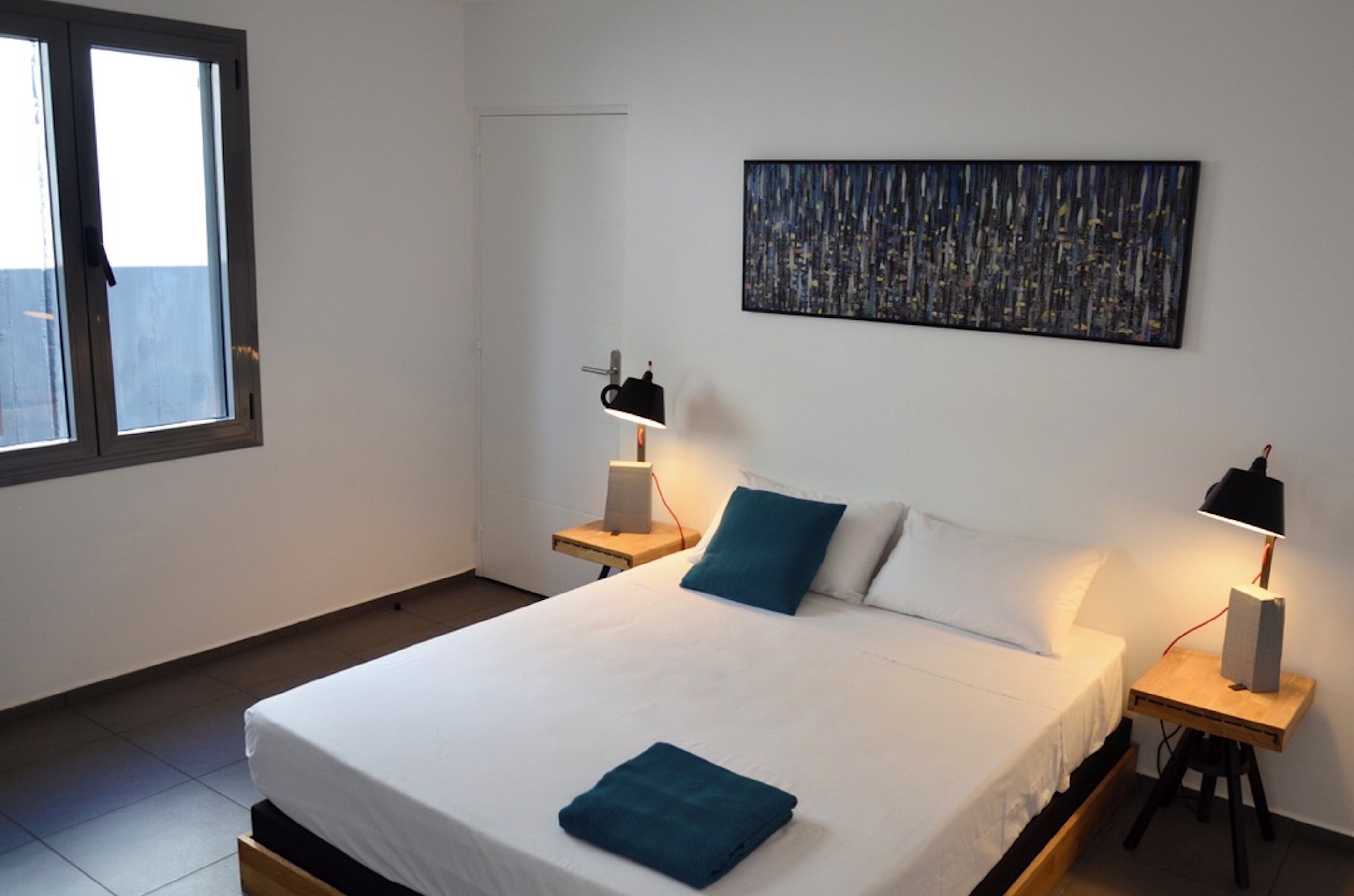 Una cama bien hecha con una toalla azul, dos lámparas de noche, una ventana y un cuadro de un paisaje urbano en la pared.