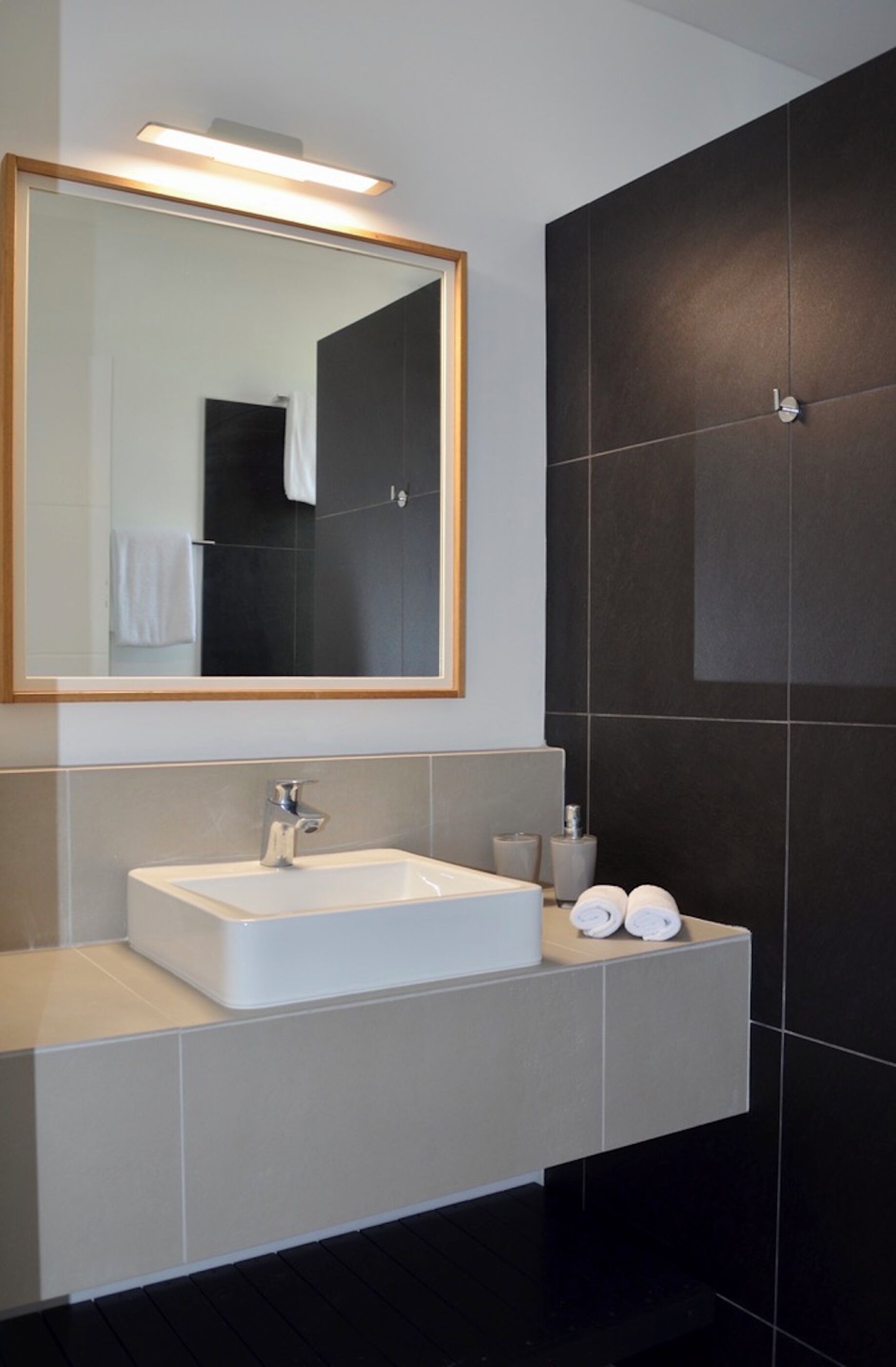 Une salle de bain moderne avec un grand miroir, un lavabo blanc et des murs recouverts de carreaux foncés.